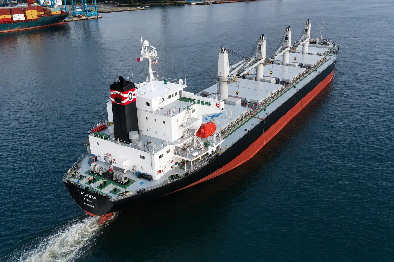 Geared handysize bulk carrier with four deck cranes under way off a container terminal, seen from above.