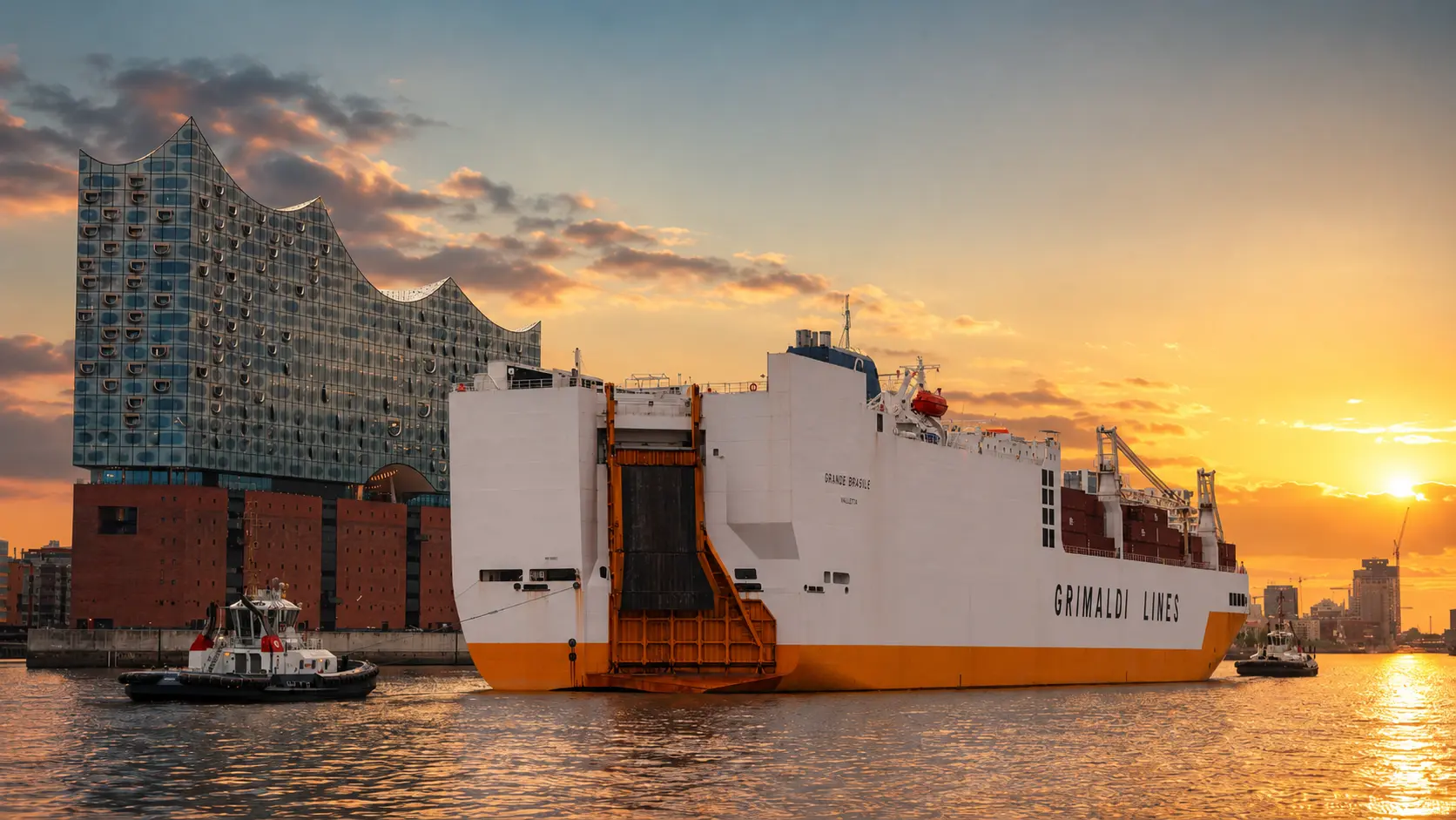 Car carrier under tug assistance in Hamburg harbour at sunset, stern ramp lowered, Elbphilharmonie visible across the water.