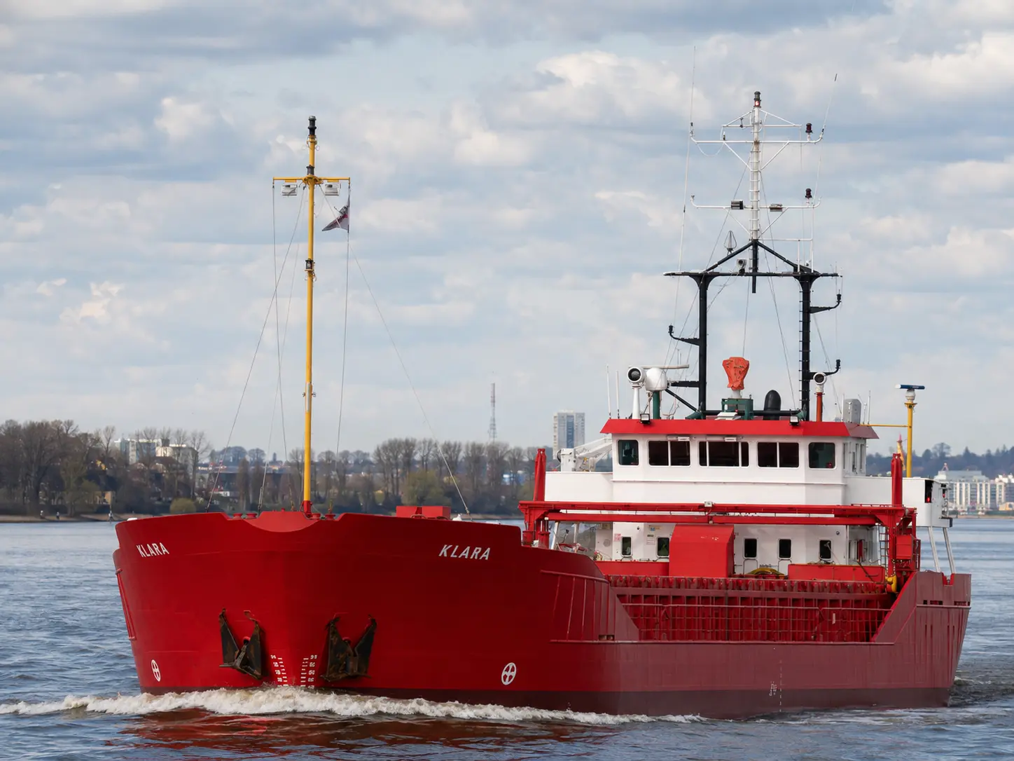Shortsea general cargo vessel KLARA with a red hull under way along a wooded shoreline, wheelhouse and accommodation block aft with the cargo hold forward.