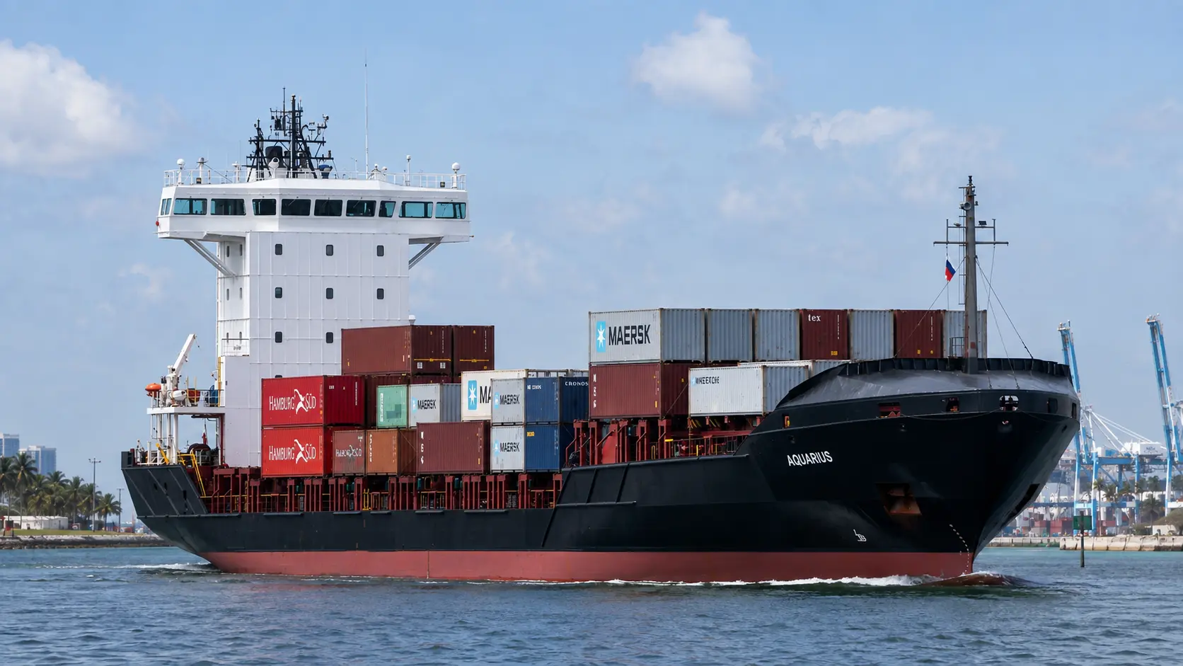 Feeder containership AQUARIUS loaded with mixed containers departing port, harbour cranes on the skyline behind.